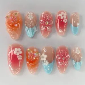 Coral sunset press on nails with iridescent aurora gold butterfly charm, 3D white floral clusters, large transparent fuchsia floral bloom with gold rhinestone center, white line art botanical illustrations on coral-red gradient base, translucent shell fan relief, aqua blue French tip accent, pearl beads, and bubble gel droplets on summer fake nails
