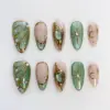 Emerald gold press on nails with 3D molten gold dragon claw frame holding green oval gemstone, sage green shimmer wing nail with gold veining and crystal rhinestones, gold star burst and crescent moon line art on nude base, and deep emerald glitter shimmer on summer fake nails