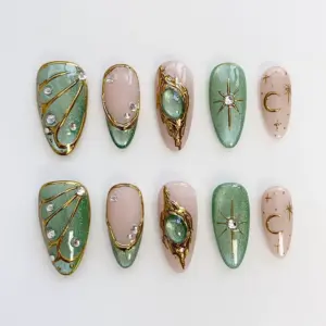 Emerald gold press on nails with 3D molten gold dragon claw frame holding green oval gemstone, sage green shimmer wing nail with gold veining and crystal rhinestones, gold star burst and crescent moon line art on nude base, and deep emerald glitter shimmer on summer fake nails