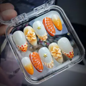 Citrus summer press on nails in a clear nail box featuring 3D realistic orange slice with water droplets, 3D peach plumeria flower with crystal bead, gold starfish charm, polka dot French tip in orange and red, and iridescent shell-pattern nail in champagne and peach fake nails