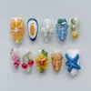 Maximalist summer art press on nails featuring 3D orange citrus slice, gold fish in navy frame, white floral bloom, blue-grey conch shell, red cherries, yellow hibiscus with green leaves, amber conch shell with pearl beads, cobalt blue starfish on white sand texture, and pink fuchsia conch with chain accent on artistic summer fake nails
