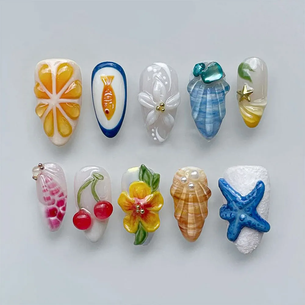 Maximalist summer art press on nails featuring 3D orange citrus slice, gold fish in navy frame, white floral bloom, blue-grey conch shell, red cherries, yellow hibiscus with green leaves, amber conch shell with pearl beads, cobalt blue starfish on white sand texture, and pink fuchsia conch with chain accent on artistic summer fake nails