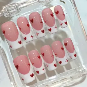 Soft pink French tip press on nails with red mini hearts and four-pointed star sparkle accents displayed in a clear acrylic nail box