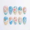 Sandy shore press on nails with hand-painted ocean wave foam on blush-pink and sky blue ombré base, 3D pink cherry blossom, lavender scallop shell, white ribbon bow, gold starfish charms, pearl micro-beads, and crystal rhinestones on summer fake nails