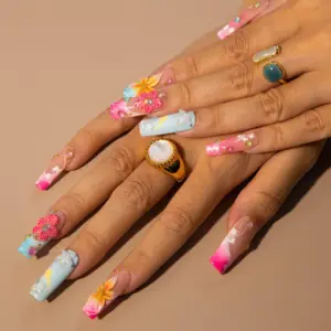 Vibrant tropical press on nails with 3D yellow sunflower and pink polka-dot flowers, white hibiscus hand-painted art, rhinestones, and pink-blue ombré on long fake nails in a clear nail box